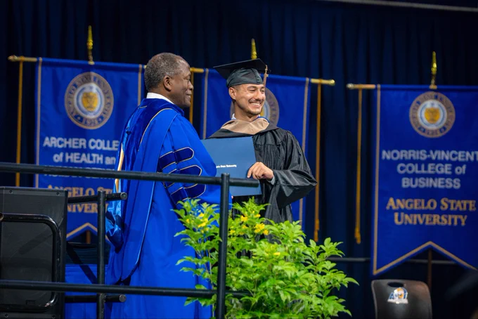 Two individuals pose for a photo together at a commencement ceremony. They’re in full graduation regalia and hold a diploma between them.