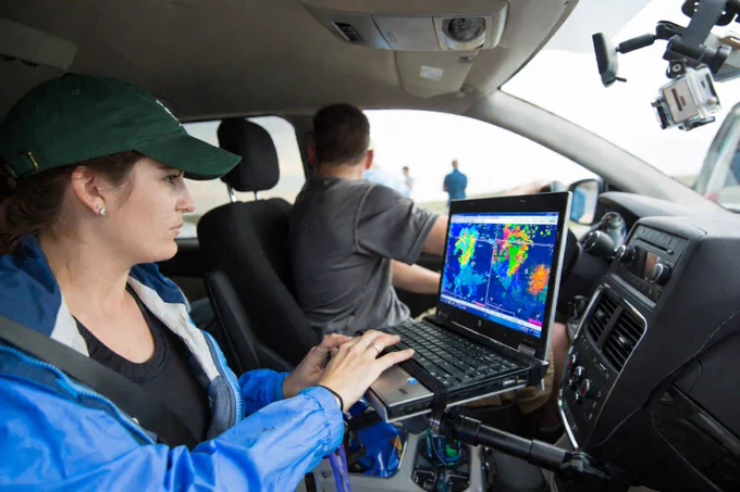 A person in a car uses a laptop displaying weather maps, with another person driving. The car is equipped with a dashboard camera. Background shows open, overcast landscape.