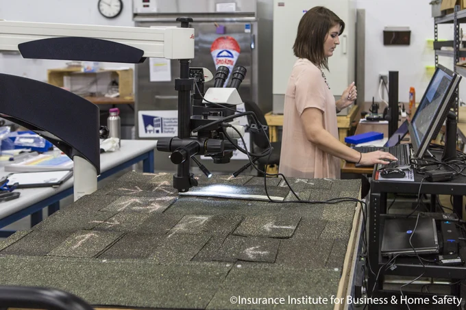 A laboratory setting showing a person working on a computer. In the foreground, there is a table with a microscope and various samples of roof shingles. The background includes equipment and a cabinet with a sign labeled "Insurance Institute for Business & Home Safety.