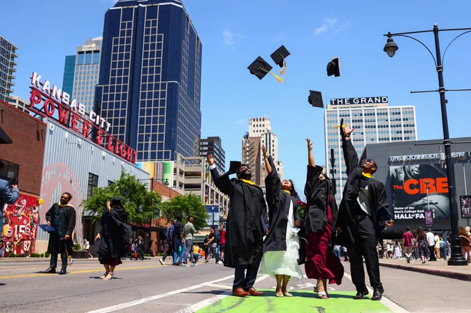 Four graduates stand in the street in downtown Kansas City and toss their caps in the air.