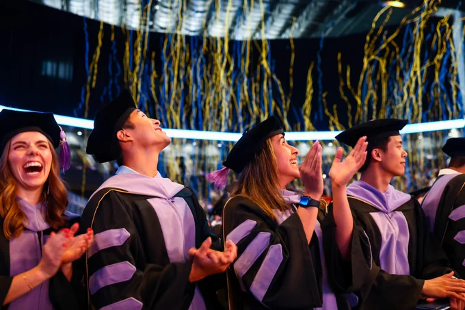 Four graduates clap and cheer as confetti falls from above them.