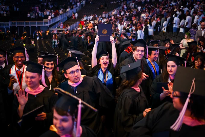 A crowd of graduates after the ceremony. One person in the middle holds up her diploma and cheers.