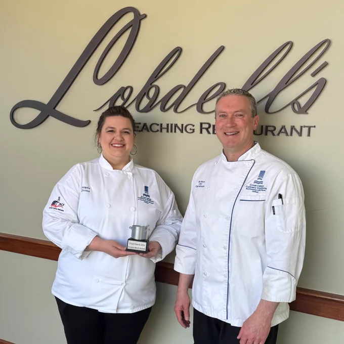 Two chefs in white uniforms stand smiling in front of a sign for Lobdell's Teaching Restaurant. One chef is holding an award.