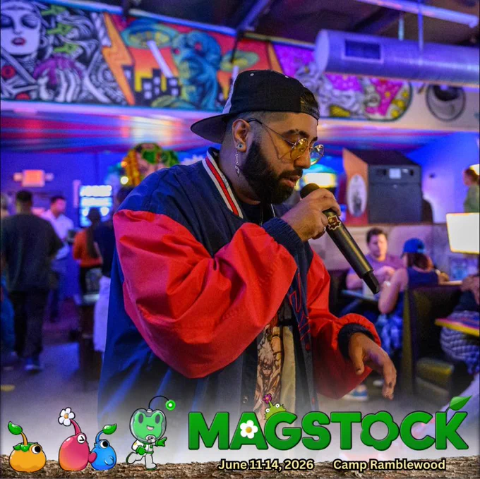 A photo of featured performer, Lord Xzae. He stands amidst a room with a scattered crowd. Some seated, others walking. Xzae is mid performance with a mic in his hand and a focus on lyrical delivery.