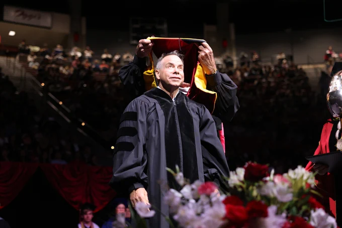 A person in academic robes is receiving a hood during a graduation ceremony, with an audience in the background. The setting includes red drapery and a table with a floral arrangement in the foreground.