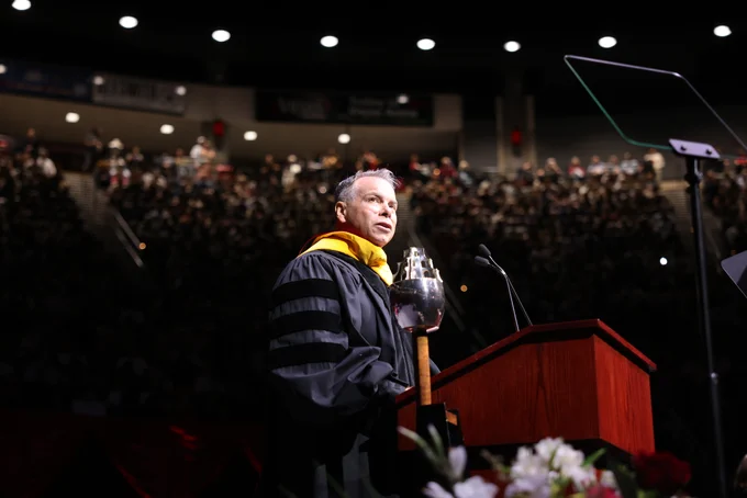 A person wearing academic regalia stands at a podium decorated with flowers, speaking to a large audience in a dimly lit auditorium. The setting suggests a formal event, possibly a graduation ceremony.