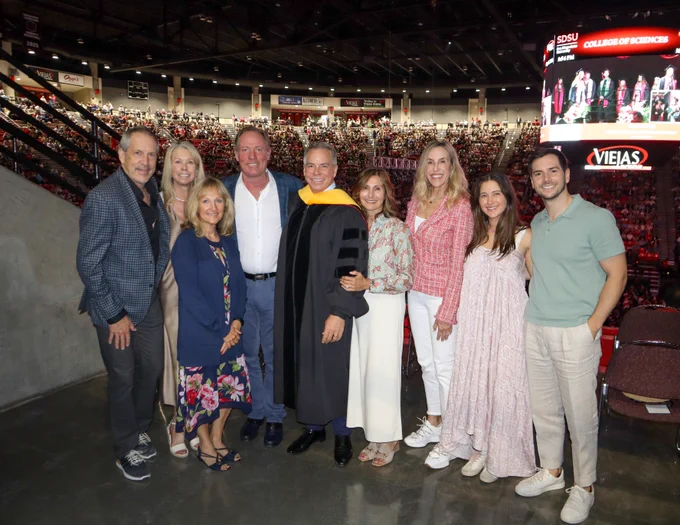 A group of people posing together in an indoor arena during a graduation ceremony. They are dressed in formal and semi-formal attire. In the background, a large crowd is seated, and a screen displays details of the event, including "Viejas Arena" and "SDSU College of Sciences".