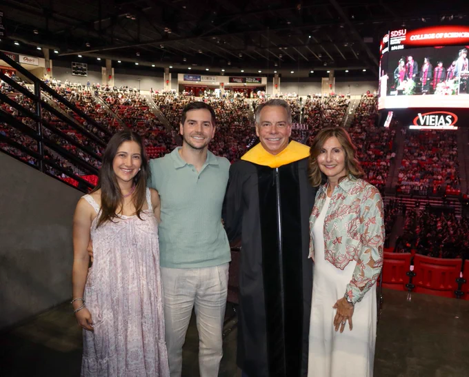 A group of four people stand together at an indoor graduation ceremony. The venue is filled with attendees and features a large screen displaying the event.