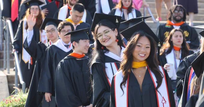 A diverse group of graduates in academic regalia walk down an outdoor staircase. Some wear stoles with Greek letters, representing fraternities or sororities. There are many attendees and graduates in the background. The atmosphere is celebratory.