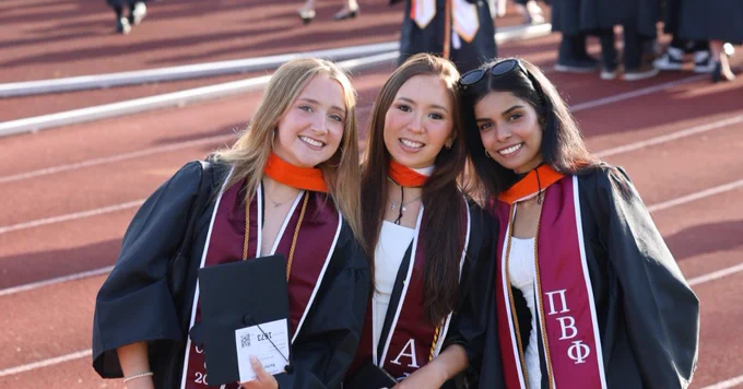 A group of graduates in caps and gowns pose for a photo on a track field. Many other graduates and attendees are visible in the background.