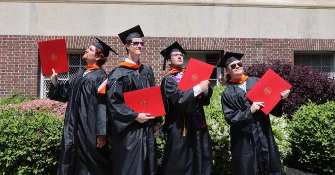 Four individuals in graduation attire are joyfully holding diplomas while posing in front of a brick building. They wear caps and gowns, standing on a sidewalk with greenery and flowers in the background.