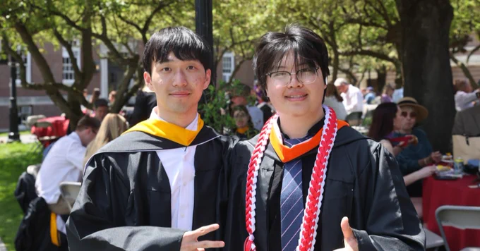 Two individuals in graduation attire stand outdoors in front of a sign for Rensselaer Polytechnic Institute (RPI). The area is grassy with tables and people in the background, and a tree provides shade.