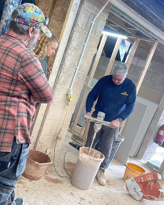 Man operating a power mixer blending construction material in a workshop while two people observe nearby.