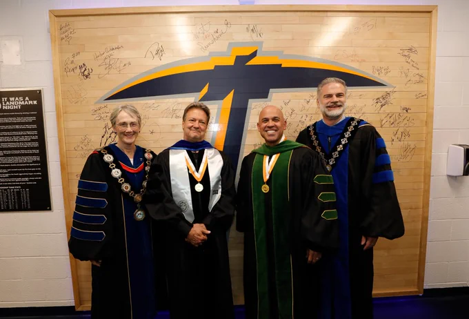 Four people wearing academic regalia stand in front of a wall featuring a large University of Texas at El Paso logo with numerous signatures.
