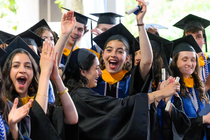 A group of graduates in caps and gowns are celebrating with smiles and raised hands. They appear joyful and excited, holding diplomas, in an outdoor setting.