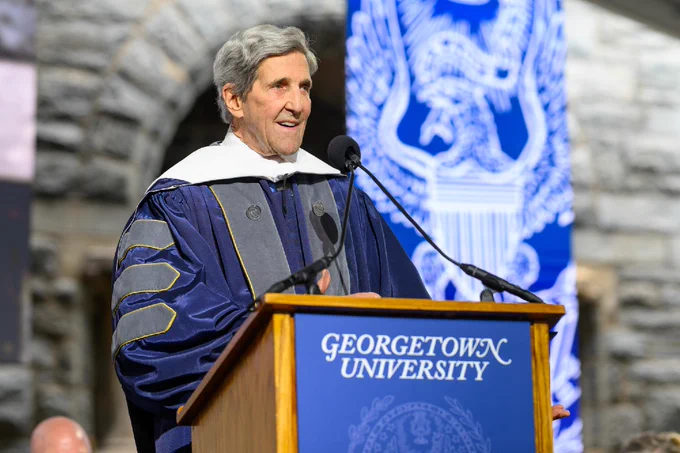 John Kerry is speaking at a podium with "Georgetown University" written on it. The background features a university emblem. The speaker is wearing academic robes.