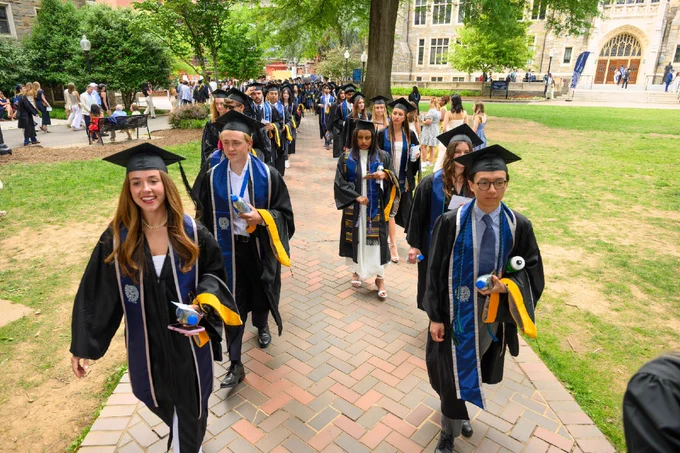 Students in graduation attire walk in a procession on a brick pathway at a university campus, with trees and buildings in the background.
