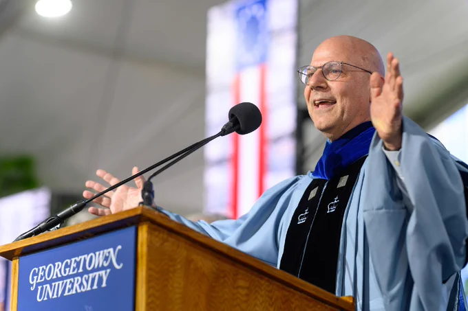 Dean Joel Hellman stands at the podium with his hands in welcome.
