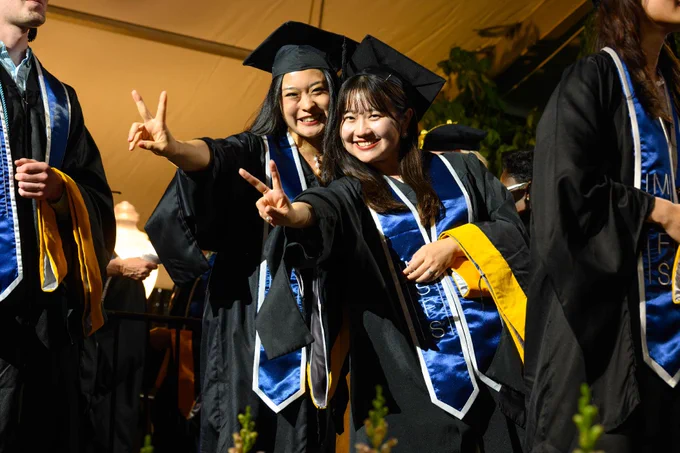 Graduates in caps and gowns pose happily, wearing blue and gold stoles. They are making peace signs at a graduation ceremony.