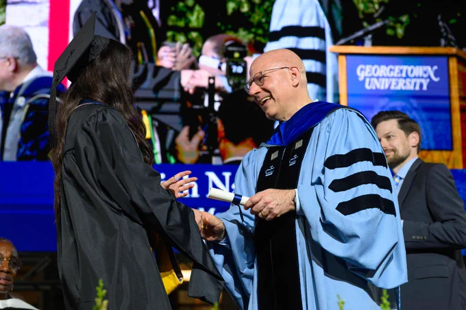 A graduating student receives a diploma translation from Dean Joel Hellman during a Georgetown University graduation ceremony. A large screen and university sign are visible in the background.