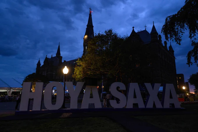 A large "HOYA SAXA" sign is displayed in front of an illuminated, historic university building at dusk. The building is surrounded by trees, with a cloudy evening sky in the background.
