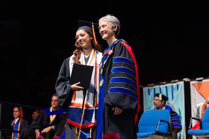 UTEP graduate Anjelica Rodriguez is in a cap and gown standing beside UTEP President Heather Wilson in regalia on a stage during a UTEP Commencement ceremony. They smile while posing for a photo, with colorful banners in the background.

