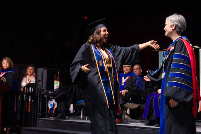 An individual in graduation attire joyfully approaches UTEP President Heather Wilson on stage during a graduation ceremony. Several seated individuals in similar attire are in the background.
