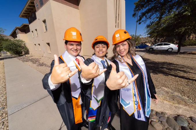 Three people are wearing orange helmets and graduation attire, including sashes and stoles, standing outdoors near a building. They are making "picks up" gestures with thumbs and pinkies extended, smiling at the camera.
