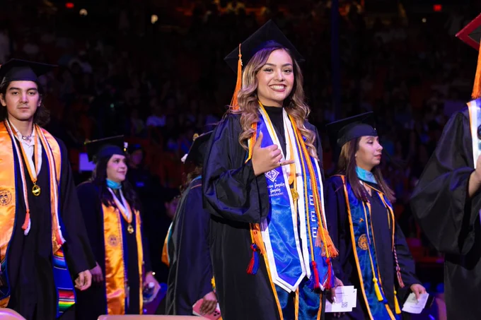 Graduates in ceremonial attire, including caps and gowns, walk in a procession during UTEP's Commencement ceremony. One individual in the foreground smiles warmly, wearing a stole with "IEEE" on it. Other graduates are visible in the background with colorful stoles and cords.