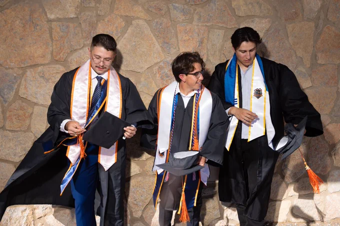 Three people in graduation attire stand in front of a textured stone wall. They are holding their caps, wearing gowns and stoles, and appear to be engaging pleasantly with each other.
