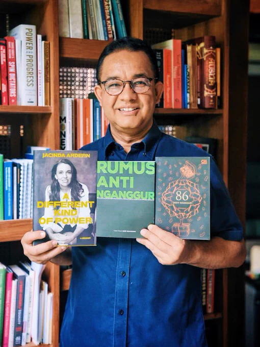 Anies Baswedan berkemeja biru tersenyum di depan rak buku kayu sambil menunjukkan tiga buku: A Different Kind of Power karya Jacinda Ardern, Rumus Anti Nganggur karya Rene Suhardono, dan 86 karya Okky Madasari.