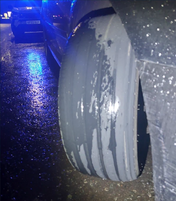 Close-up of a severely worn and bald car tyre on a wet road at night, with blue police lights reflecting.