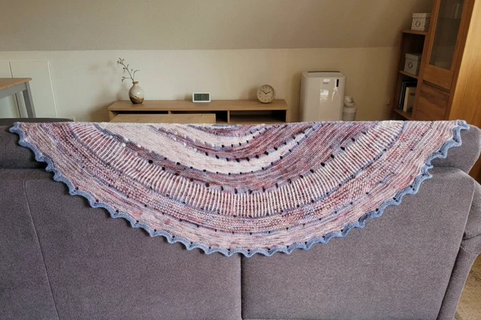 Ein detailreiches Stricktuch in Form eines großen Halbkreises hängt ausgebreitet über der Rückenlehne eines grauen Sofas. Die Decke ist in harmonischen Mustern gestrickt und zeigt konzentrische Kreise in sanften Violett-, Rosa-, Creme- und Pastelltönen mit kleinen Durchbruchmustern (Ajour-Elementen) und einem welligen, hellblauen Abschlussrand.
Das Sofa steht mittig im Bild.
Der Hintergrund ist ein ordentliches, helles Wohnzimmer. Hinter dem Sofa steht ein niedriges Holzregal, auf dem eine schlichte Blumenvase, eine kleine Digitaluhr und eine analoge Tischuhr stehen. Rechts im Hintergrund sind ein weißes Standgerät und ein heller Holzschrank zu sehen, links ein Teil eines Esstisches. Das Licht ist gleichmäßig, hell und freundlich, ohne harte Schatten.