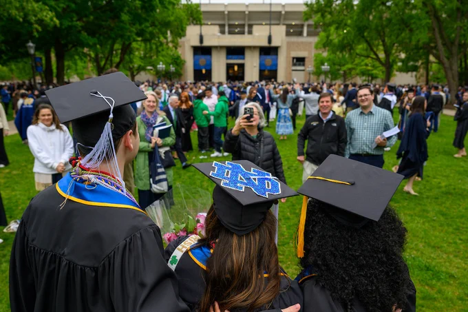 A group of three individuals in graduation caps and gowns are posing for a photo. The center person's cap features a University of Notre Dame emblem. They stand amidst a crowd on a green lawn with a large building in the background.