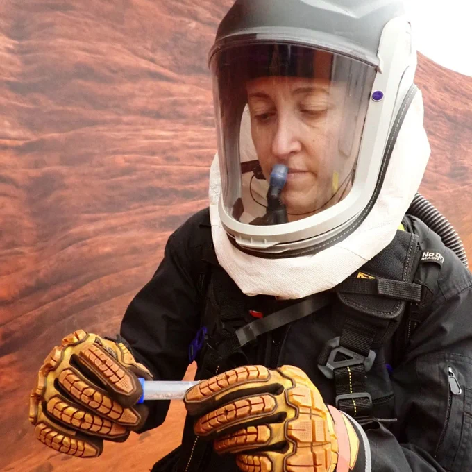 CHAPEA mission 2 medical officer Ellen Ellis wearing a protective helmet and gloves holding a small blue and white object against a textured reddish rock background. She collects samples during an extravehicular activity. (c) NASA