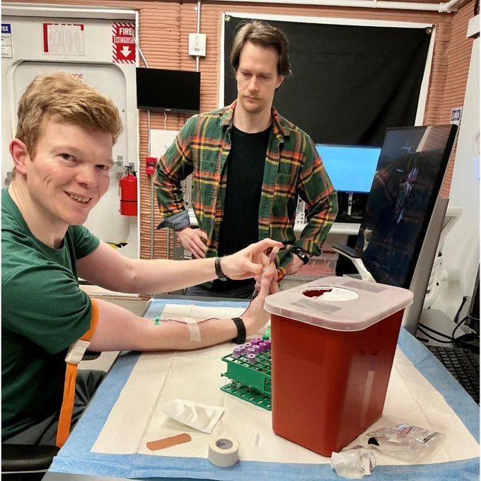 CHAPEA crew member James Spicer seated with arm extended for blood draw, Matthew Montgomery standing nearby to perform blood collections to monitor their health. (c) NASA