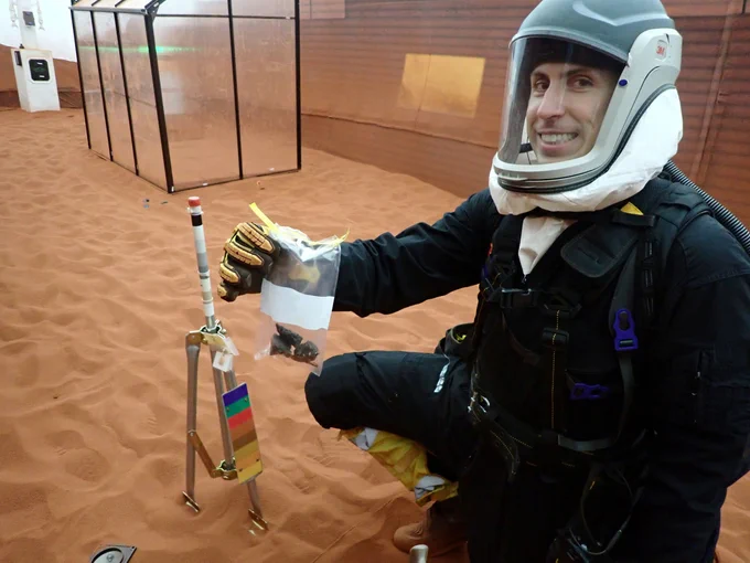 CHAPEA mission 2 commander Ross Elder in protective suit collecting geological samples inside a controlled sandy environment during a simulated extravehicular activity. (c) NASA