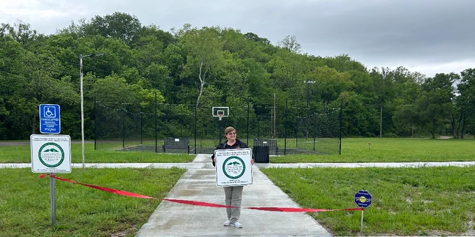 Woman standing behind a red ribbon in front of pickleball court holding a sign talking about Land and Water Conservation Fund.