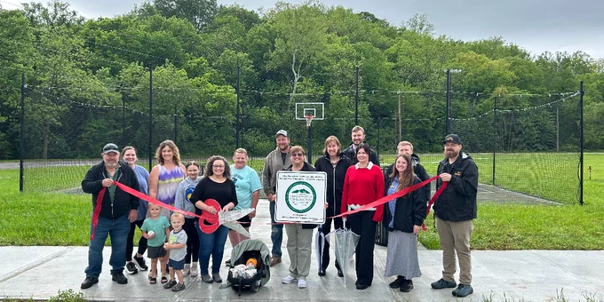 Group of people gathered outdoors on a basketball court holding a red ribbon for a ribbon-cutting ceremony with trees in the background.