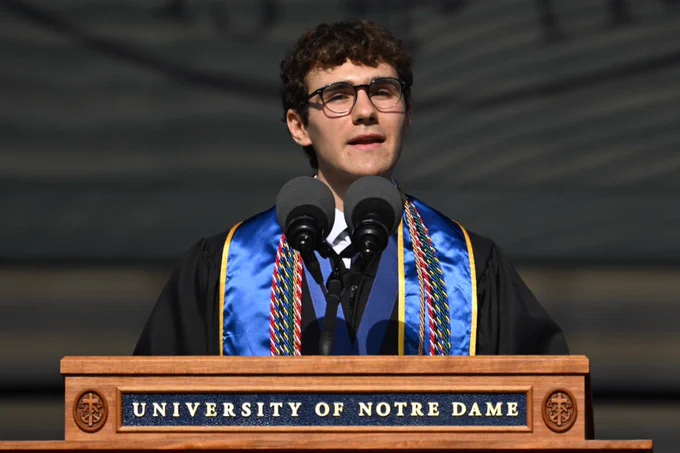 A person wearing glasses and a graduation gown stands at a podium with the words "University of Notre Dame." They are speaking into multiple microphones. The person is adorned with a blue stole and several multi-colored cords.