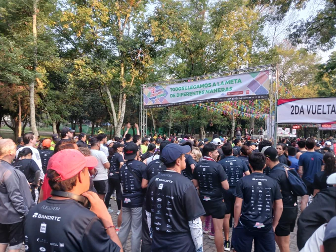 Fotografía a color al interior del Bosque de Chapultepec, en la que aparecen personas asistentes a la Carrera Corre por Ti, Camina Conmigo 2025 preparándose para la salida y en la parte superior un letrero con la frase: Todos llegamos a la meta de diferentes maneras.