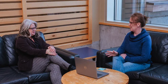 Two people sit on black leather couches in a casual setting, engaged in conversation. A wooden coffee table between them holds a Dell laptop and a smartphone. The room features wooden paneling and large windows.