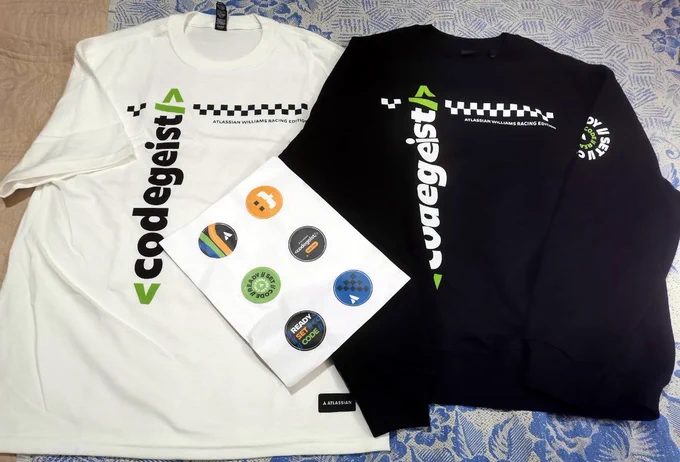 Two items of exclusive Atlassian Codegeist 2025 Williams Racing Edition developer swag: a white long-sleeve t-shirt and a black sweatshirt, both featuring the green logo and racing-style checkered patterns. A sheet of branded stickers is placed in the center.