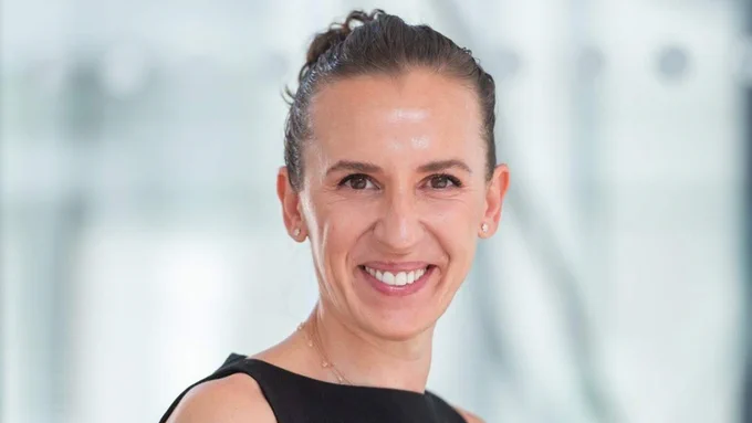 A headshot of a smiling woman with her hair pulled back, wearing a black dress, is M\u00E1rta C\u00E2rsteanu-Dombi. She is fair-skinned with brown hair and eyes, and a bright white smile. Her backdrop is blurred and light.