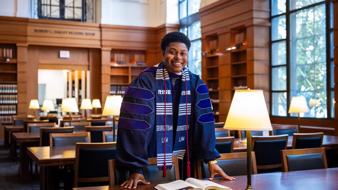A man in Juris Doctor regalia stands over a book in a library, smiling