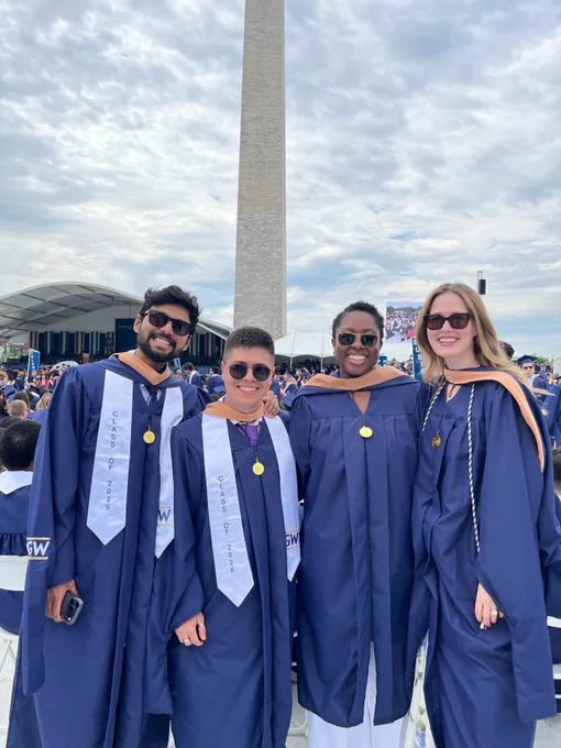 Graduates in blue robes smile in front of the Washington Monument during the commencement ceremony. A stage is visible in the background.