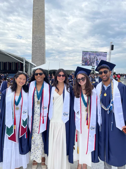 A group of graduates wearing caps and gowns stands together, smiling. They are in front of the Washington Monument, with an outdoor stage and screen visible in the background.