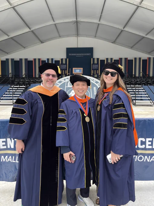 Three individuals in academic regalia, wearing caps and gowns, stand together smiling in front of a stage. The backdrop features a podium and banners.