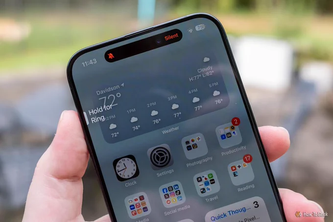 Hand holding an iPhone outdoors, pressing the side Action button while a "Silent" indicator and muted-bell icon appear in the Dynamic Island, showing the button toggling the phone's ring/silent mode.