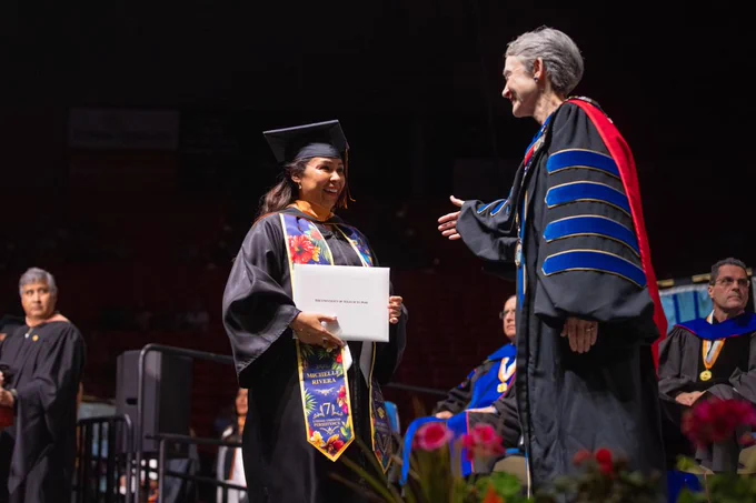 Michelle Cain, a graduate in a black cap and gown, is shaking hands with UTEP President Heather Wilson, dressed in academic regalia, at a UTEP commencement ceremony. A diploma is held in one hand, and a decorated stole is worn. Other officials are visible in the background on stage. Vibrant flowers are in the foreground.
